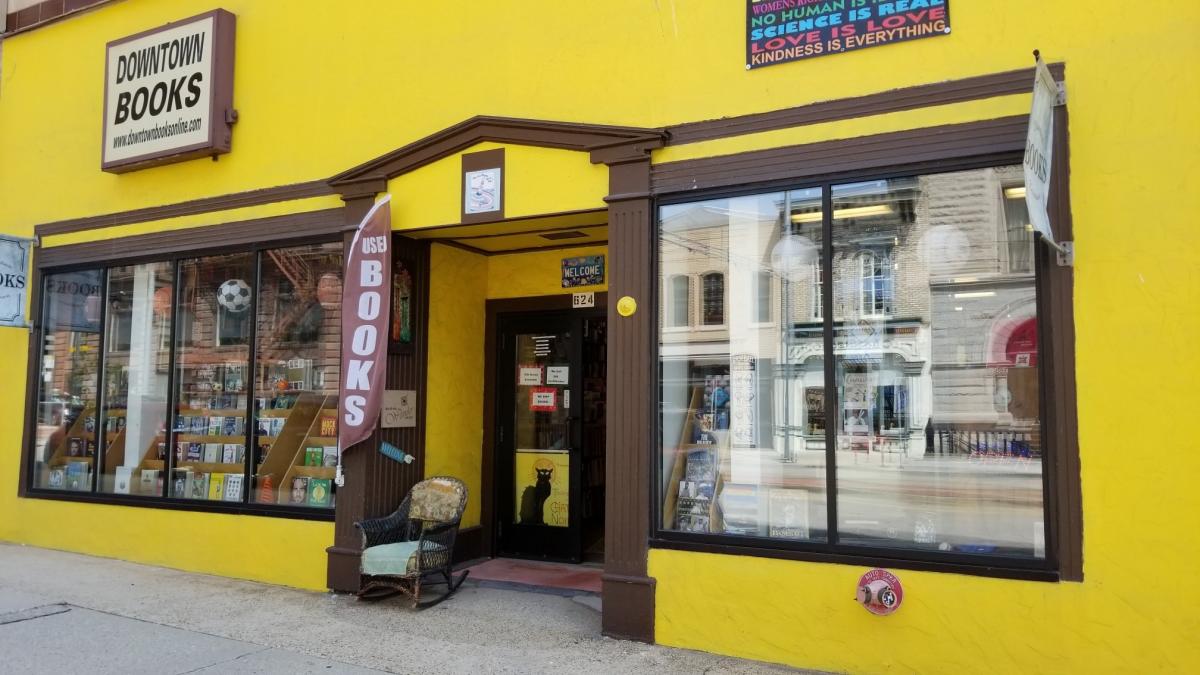 Exterior of Downtown Books, a used bookstore with a bright yellow facade, large front windows displaying books, a brown “Used Books” flag by the entrance, and a wicker chair outside.