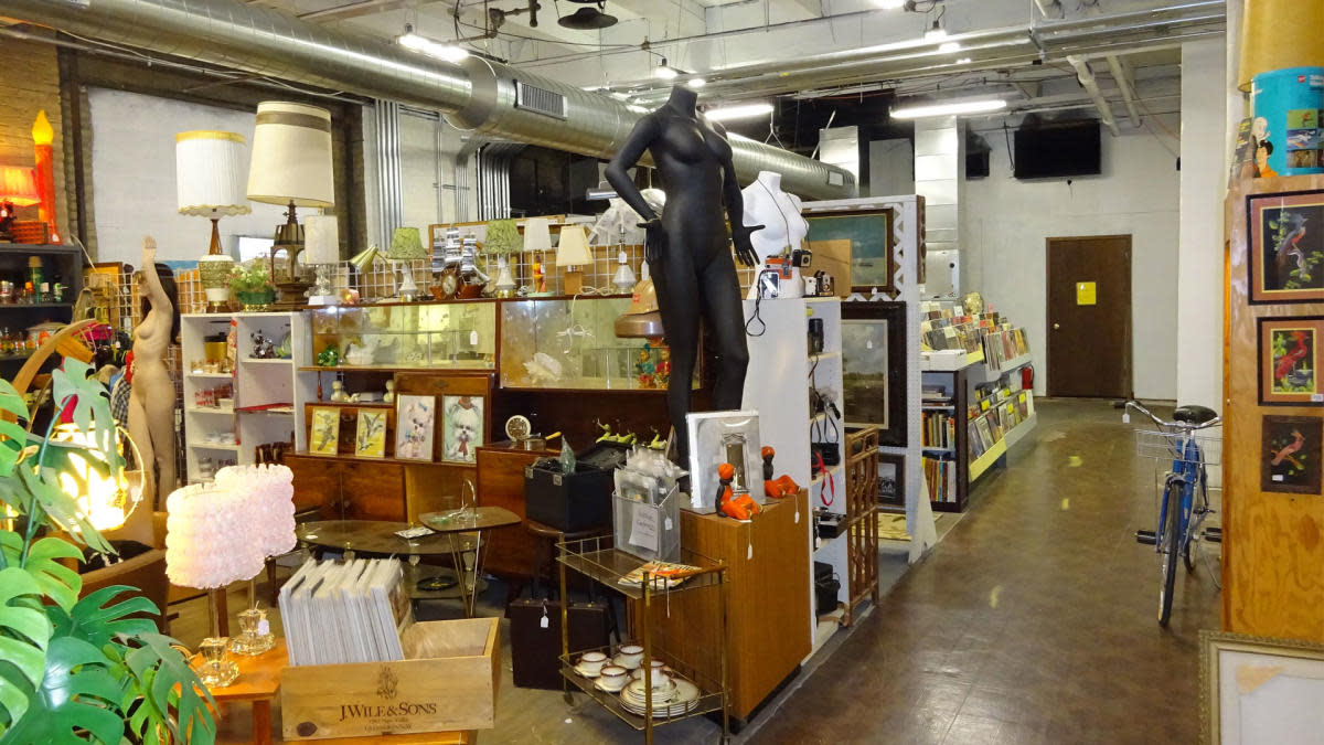 A showroom inside Antiques on Pierce displays an eclectic collection of vintage furniture, lighting, artwork, records and home décor. Glass display cases, mid-century furnishings, mannequins and collectibles fill the industrial-style space, with wide aisles inviting shoppers to browse.