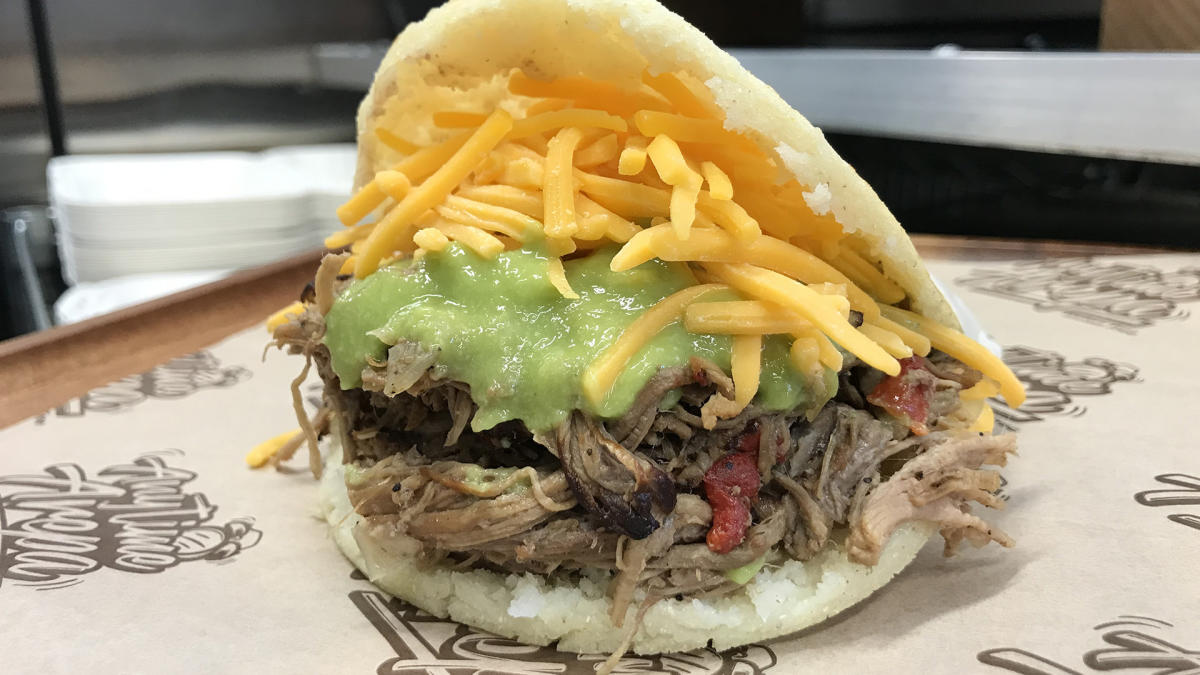 A close-up of a loaded arepa from Anytime Arepa at American Family Field. The sandwich is packed with shredded beef, melted cheddar cheese, and green sauce, spilling out of a warm, golden cornmeal pocket.