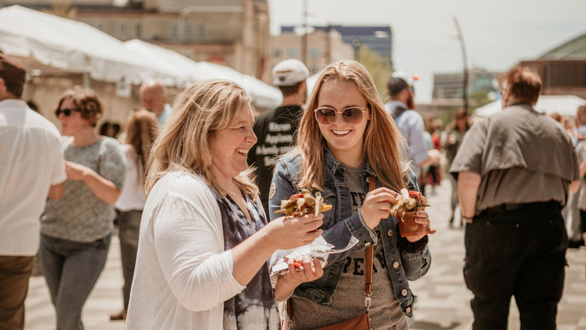 Two women smile and enjoy elaborately garnished Bloody Marys at an outdoor festival, surrounded by other attendees and white vendor tents under a sunny sky in an urban setting.