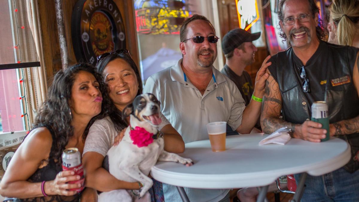 Four people gather around a small round table inside a bar, smiling and holding drinks. A small dog with a pink flower collar sits on one person’s lap, tongue out happily. Neon signs and a Lakefront Brewery sign are visible in the background, adding to the lively atmosphere.