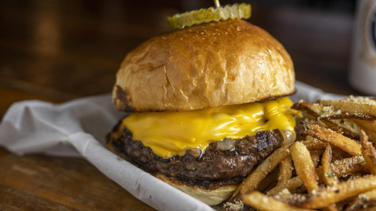 Cheeseburger with a thick beef patty and melted cheddar on a toasted bun, topped with sliced pickles and served with seasoned fries on a parchment-lined tray.