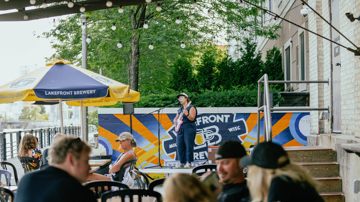 A woman in a navy blue romper singing into a microphone on a stage, holding a pink guitar, with people in front of her sitting at tables under gold and blue umbrellas on a patio along the riverfront with trees in the background.