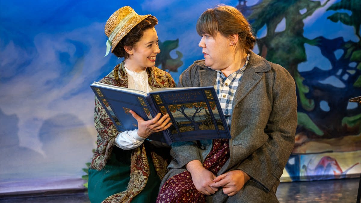 Two actors sit on a stage as one reads from a book and the other reacts with surprise.