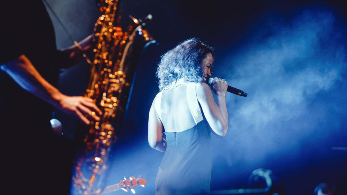 A female jazz singer performs on stage in a sleeveless black dress, holding a microphone as blue stage lights and smoke create a moody atmosphere; a saxophonist is partially visible in the background.