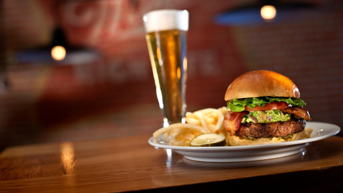 Burger topped with bacon, lettuce, tomato, and avocado on a bun, served with French fries and pickles on a white plate, with a glass of beer in the background.