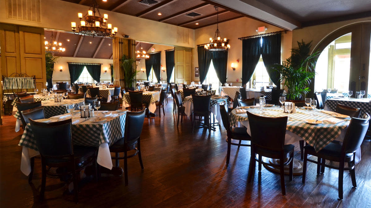 Elegant dining room with round tables covered in green and white checkered tablecloths, set with glassware and silverware under warm chandeliers, surrounded by dark wood chairs and large windows with dark drapes.