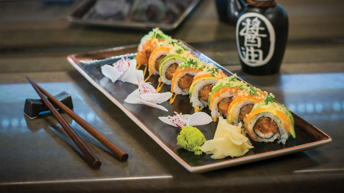 Sushi roll sliced into pieces and topped with avocado and salmon on a rectangular black plate, served with wasabi, pickled ginger, and edible flowers, with chopsticks and a soy sauce bottle nearby.