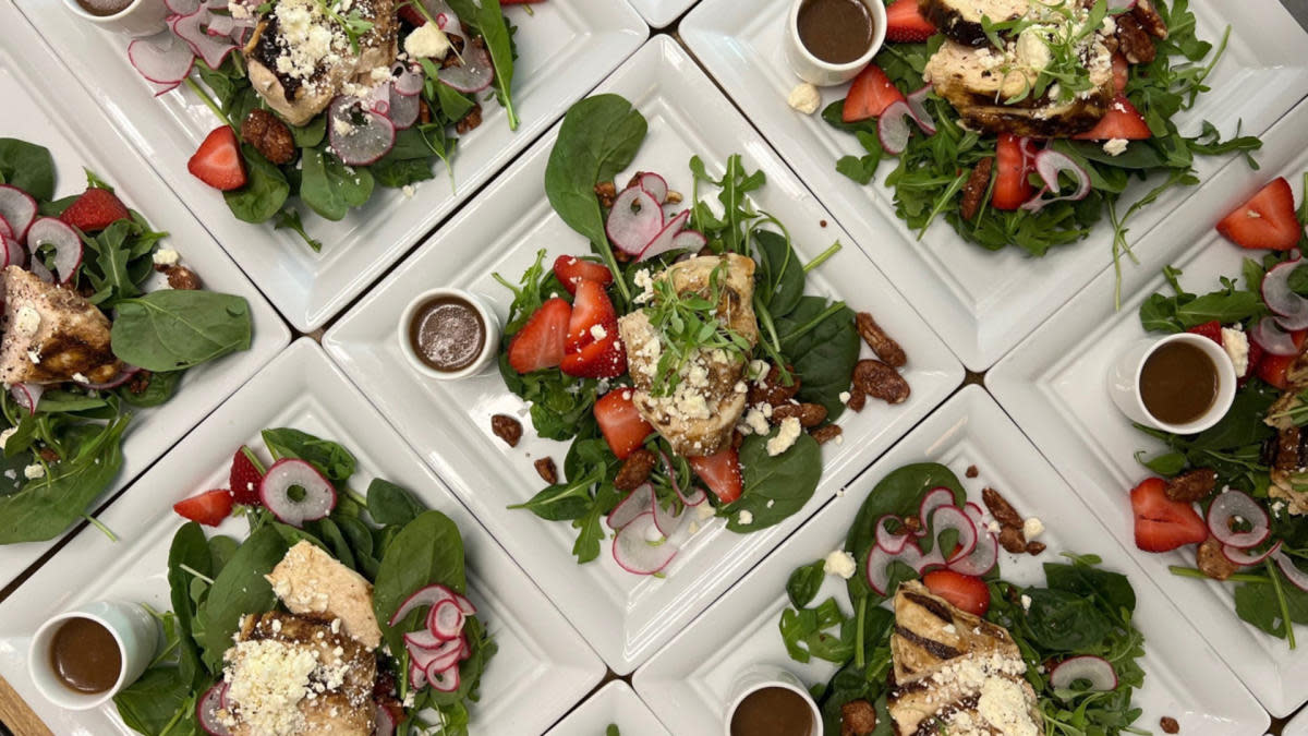 Plates of fresh salads arranged in a grid, each featuring grilled chicken breast on a bed of spinach and arugula with sliced strawberries, radishes, candied nuts, crumbled cheese, and a small cup of balsamic dressing on white square dishes.
