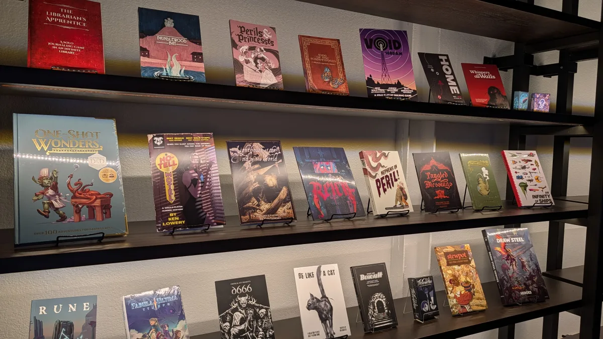 Shelving unit displaying a curated selection of tabletop and role playing game books, arranged face out on black shelves. Titles vary in size and style, with colorful illustrated covers ranging from fantasy themes to horror and indie designs. A larger book titled “One Shot Wonders” is prominently featured on the left, while smaller titles line the upper and lower shelves. Soft lighting highlights the display against a neutral wall, giving the setup a clean, gallery like presentation.
