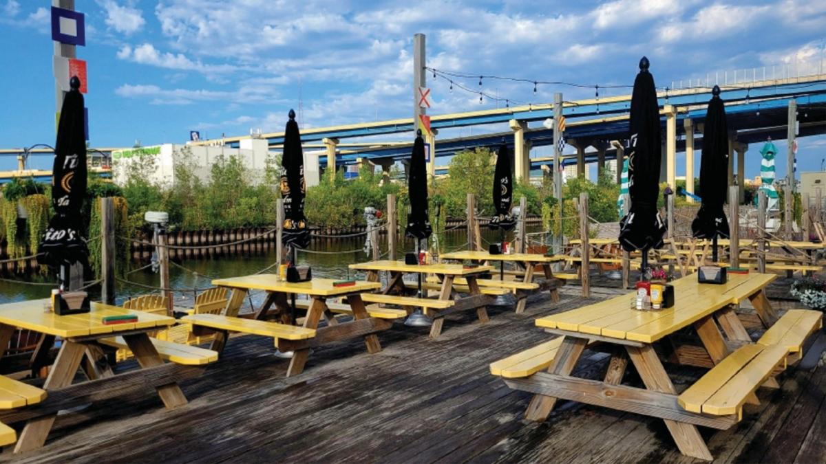 Outdoor riverside dining area with yellow picnic tables, black umbrellas, and string lights at a waterfront restaurant on a sunny day. A highway bridge and greenery are visible in the background.