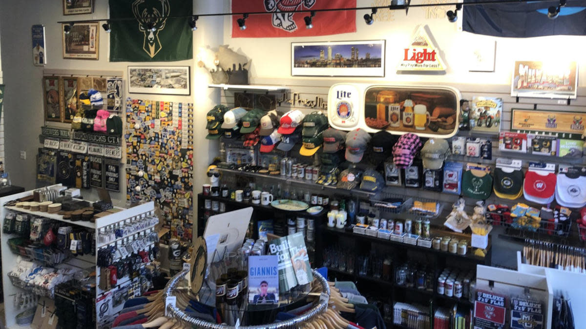 Interior of a Milwaukee-themed retail shop filled with local sports and city merchandise. Shelves and wall displays are packed with baseball caps, glassware, mugs, magnets, and souvenirs featuring teams like the Bucks and Brewers. Framed posters, vintage signs, and banners line the upper walls, while a central display table holds books and additional items. The space is brightly lit and densely arranged, creating a colorful, curated storefront focused on Milwaukee pride.
