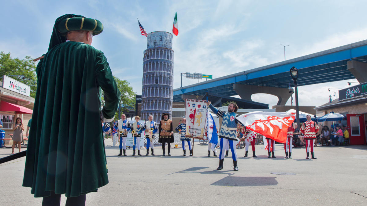 Performers in traditional Renaissance costumes participate in a flag-throwing demonstration at Milwaukee’s Festa Italiana, with a replica of the Leaning Tower of Pisa and both Italian and American flags in the background. Spectators watch from nearby food stands and shaded seating areas under a sunny sky.