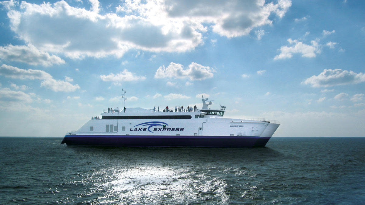 The Lake Express high-speed ferry crossing Lake Michigan on a sunny day, with passengers standing on the upper deck under a partly cloudy sky.
