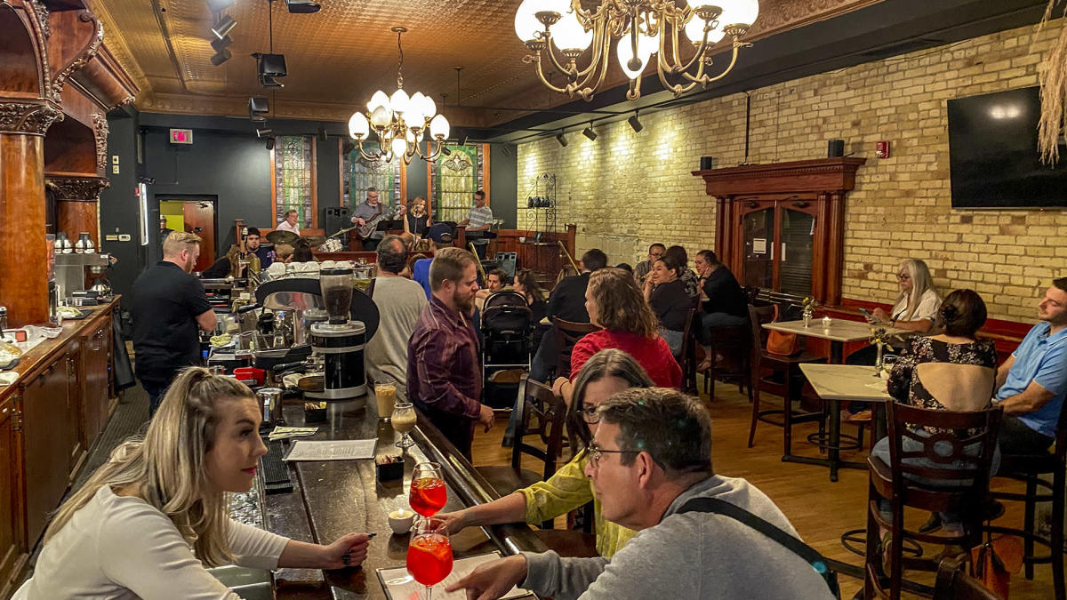 An elegant, warmly lit bar and café with ornate woodwork and chandeliers, filled with people socializing at tables and the bar. A small band performs on a raised stage at the back of the room. Patrons chat, drink, and enjoy the lively atmosphere, with exposed brick walls and vintage décor adding to the cozy ambiance.