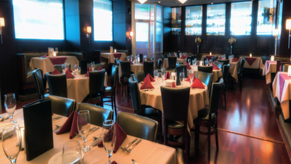 Upscale restaurant dining room with round tables covered in beige tablecloths and red folded napkins, surrounded by dark leather chairs and softly lit by wall sconces and candles, creating a warm, elegant atmosphere.