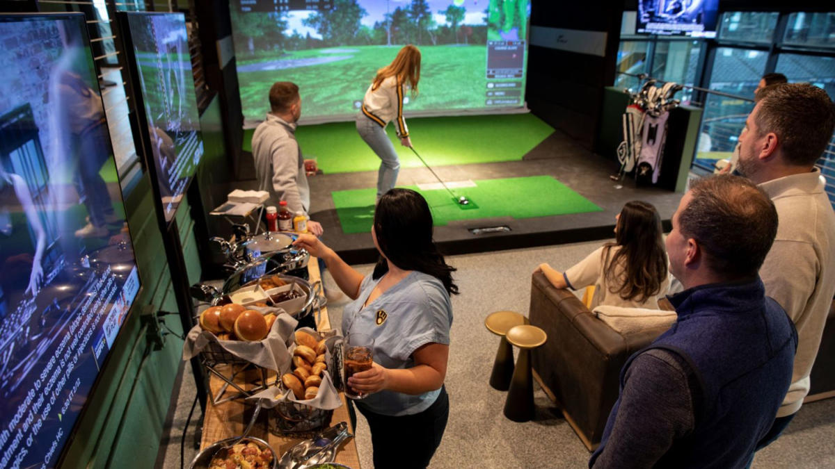 A group of people enjoy food and drinks while watching a woman play on an indoor golf simulator at X-Golf inside American Family Field. Multiple TV screens, baskets of buns, and bar-style seating create a lively social atmosphere.