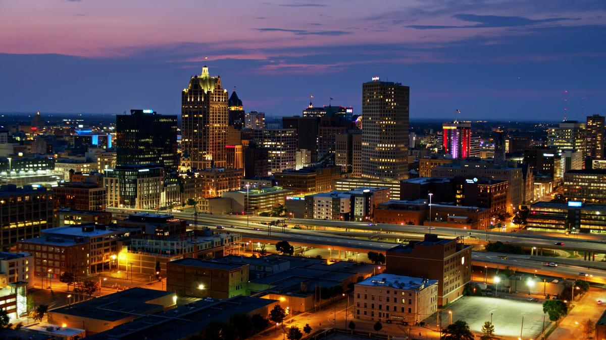 A twilight view of downtown Milwaukee, Wisconsin, featuring illuminated skyscrapers and city lights against a purple and blue sunset sky, with highways and streets visible in the foreground.