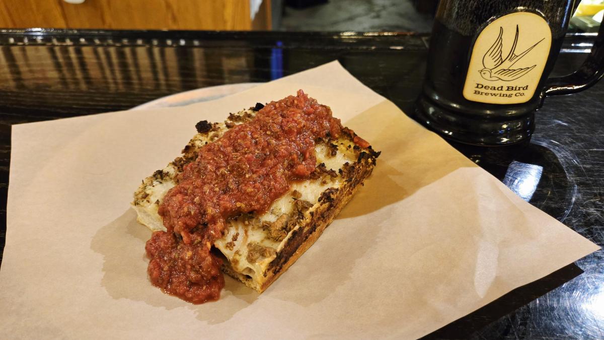 A thick slice of Detroit-style pizza topped with chunky red sauce and melted cheese, served on parchment paper beside a black Dead Bird Brewing Co. mug.