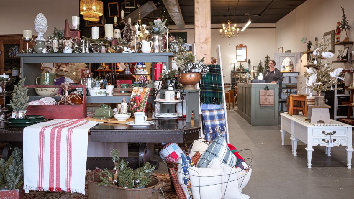 Interior of a vintage shop with assorted items like furniture, decorations, and festive ornaments displayed for sale, with a person standing by the counter in the background.