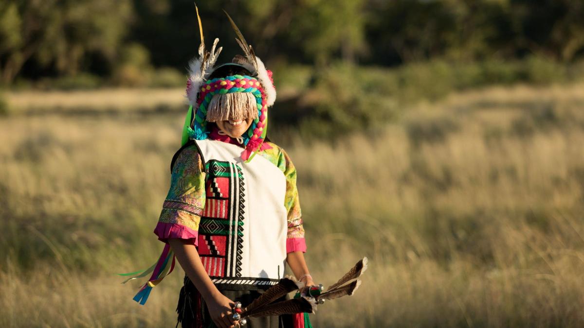 A Puebloan dancer in traditional garb stands in a grassy field.