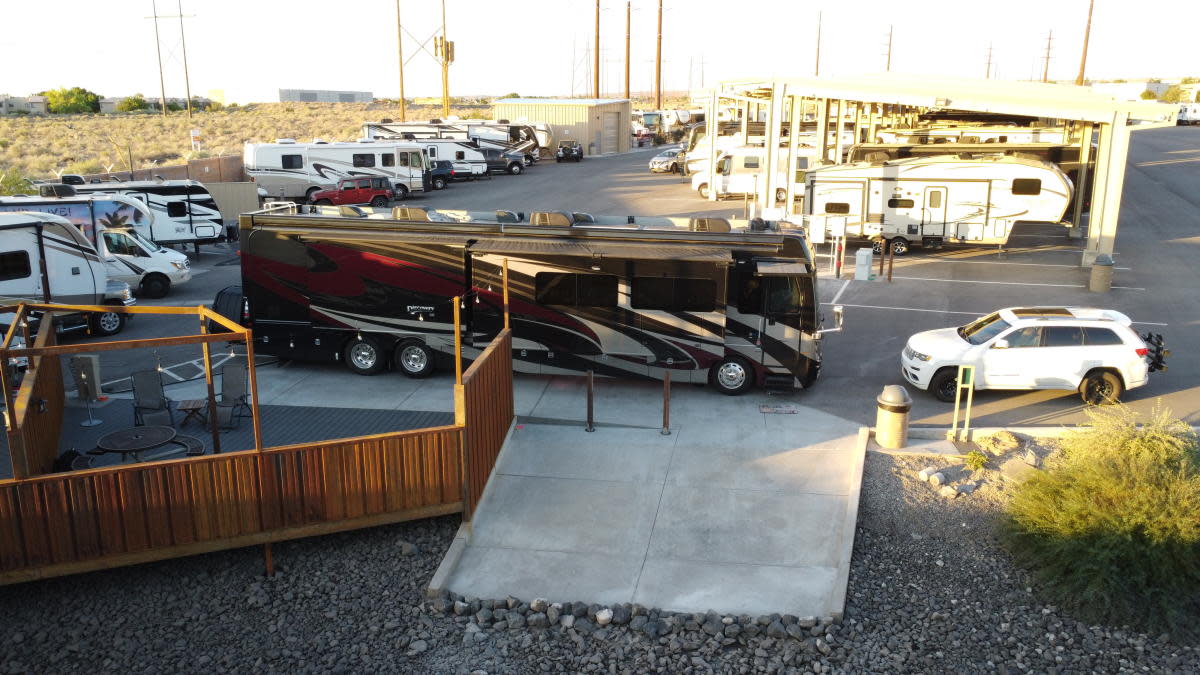A large RV sits in front of a parking lot with many smaller RVs are parked under covered awnings with telephone poles and desert plains in the background.
