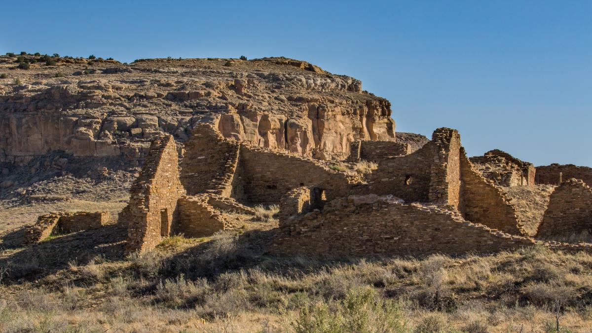 Ruins at Chaco Culture National Historical Park, showing crumbling ancient walls against a backdrop of a large cliff under a clear blue sky.