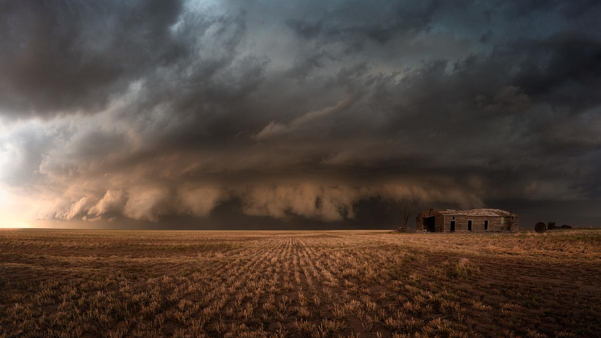 A massive, dark storm cloud hovers over an abandoned farmhouse and a barren field.