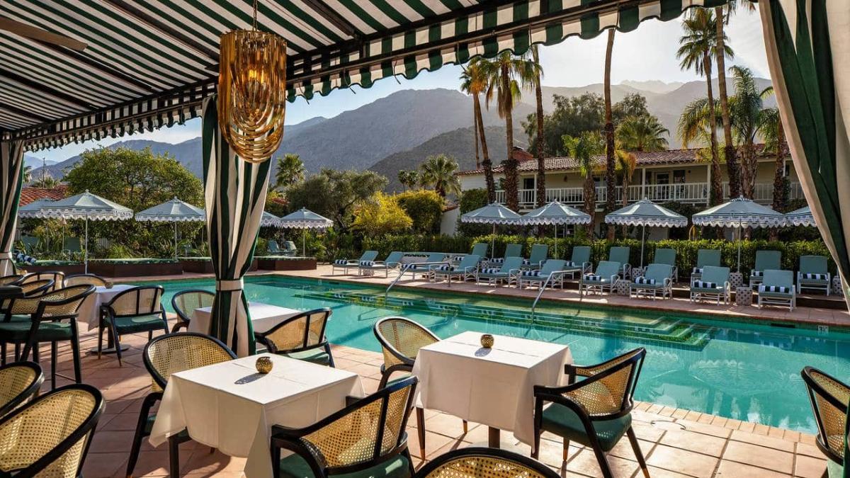 Outdoor pool area of a luxury resort with striped umbrellas and lounge chairs, surrounded by palm trees and mountains in the background, viewed from under a striped canopy with a hanging light fixture. Dining tables are set up in the foreground.