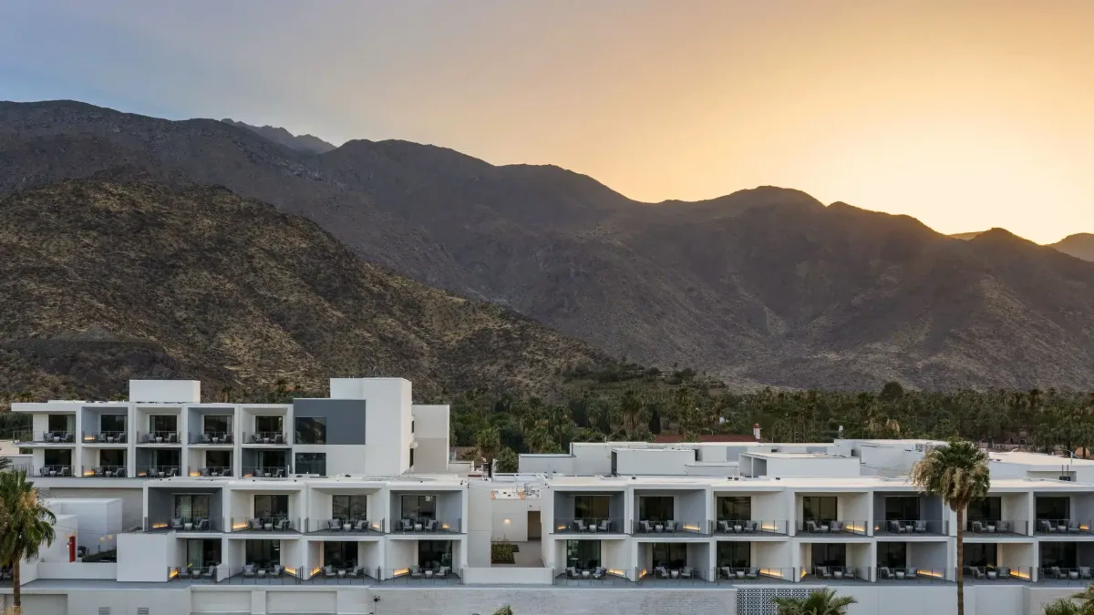 A modern multi-story building with balconies in the foreground, with a backdrop of rolling mountains under a soft sunset glow.