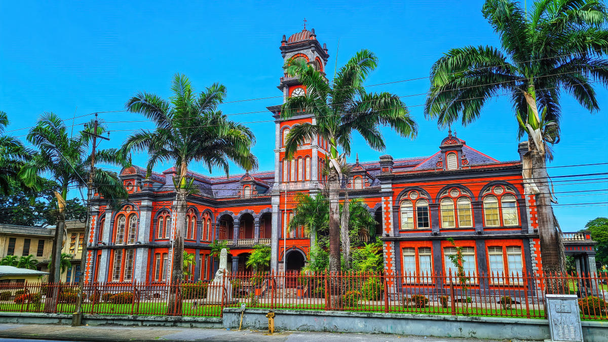 A historic brick building with a clock tower and intricate architectural details. The building is painted in shades of red and gray and is surrounded by palm trees.