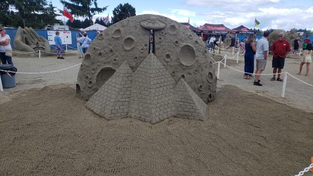 Sand sculpture depicting three pyramids in front of a large moon. Over the top of the middle pyramid is a UFO, and coming down from the UFO is a sliver of blank space.