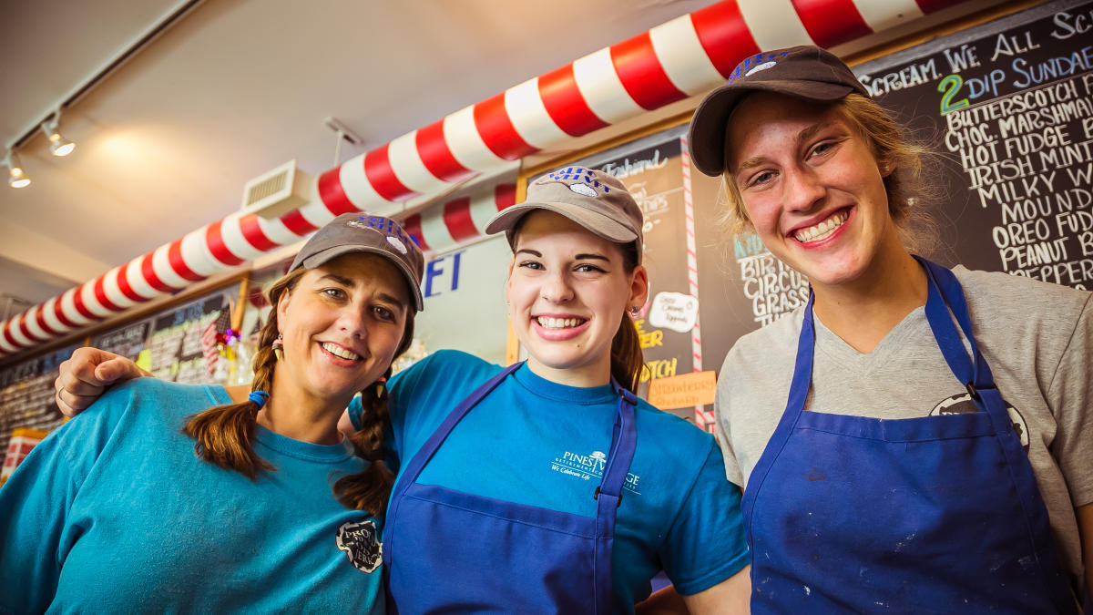 Three women smile in front of a chalkboard menu. All are wearing baseball hats.