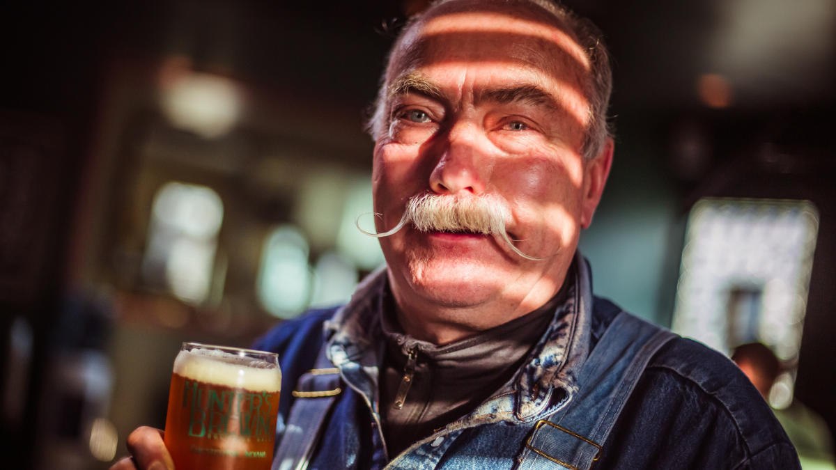 An older man with a curled mustache holds a glass of beer.