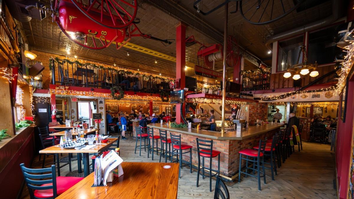 An empty restaurant is lit by a combination of natural light from the windows out of view on the left and soft white interior lights. The restaurant features red and black chairs, wooden tables, and vintage firehouse decor throughout.