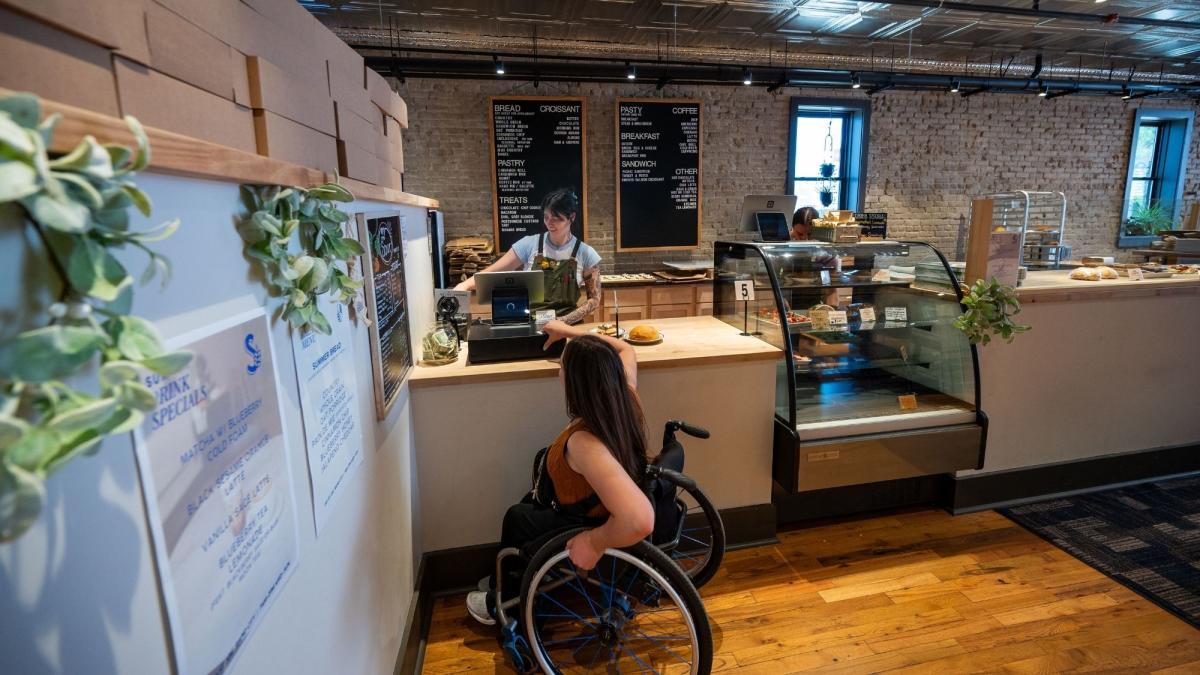A customer in a wheelchair at The Sour counter, interacting with a cafe worker. The cafe has a cozy, rustic style with wood accents and a menu board.