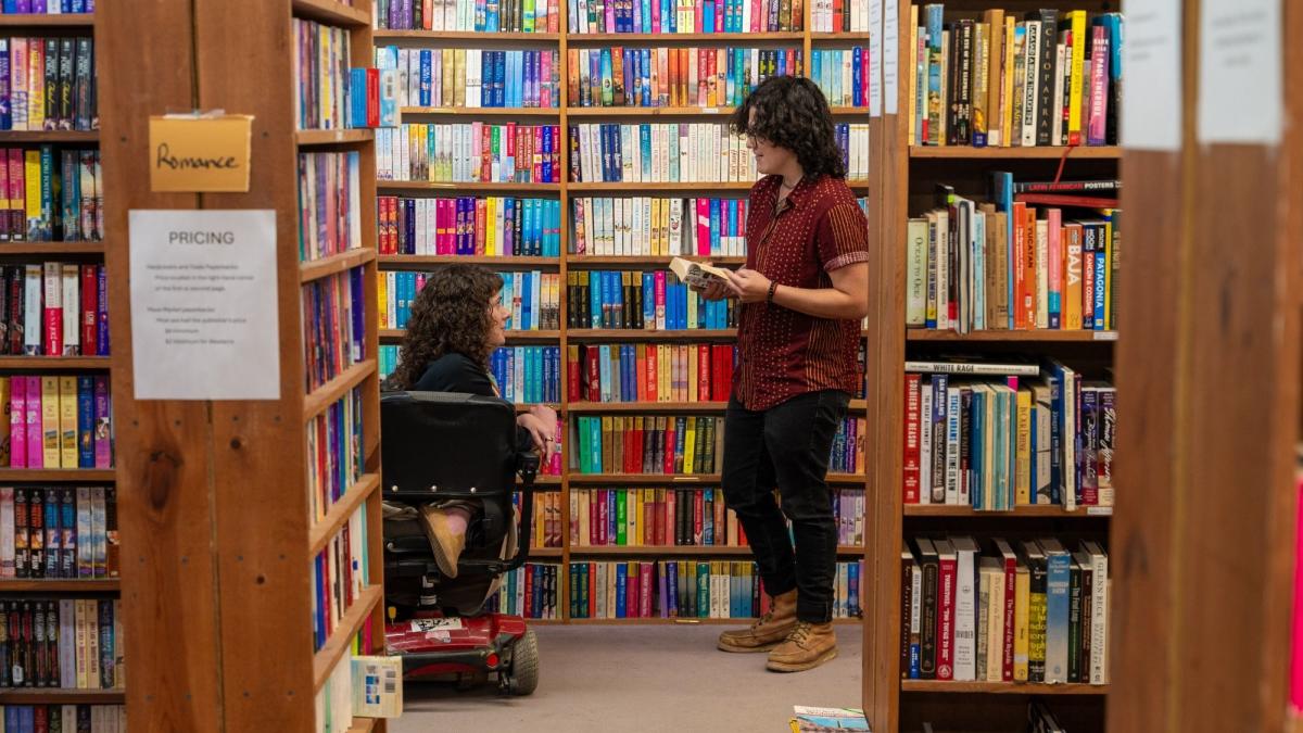Two people are in a cozy bookstore aisle lined with colorful books. One person in a wheelchair reads a book, while the other stands, also reading. The atmosphere is warm and engaging.