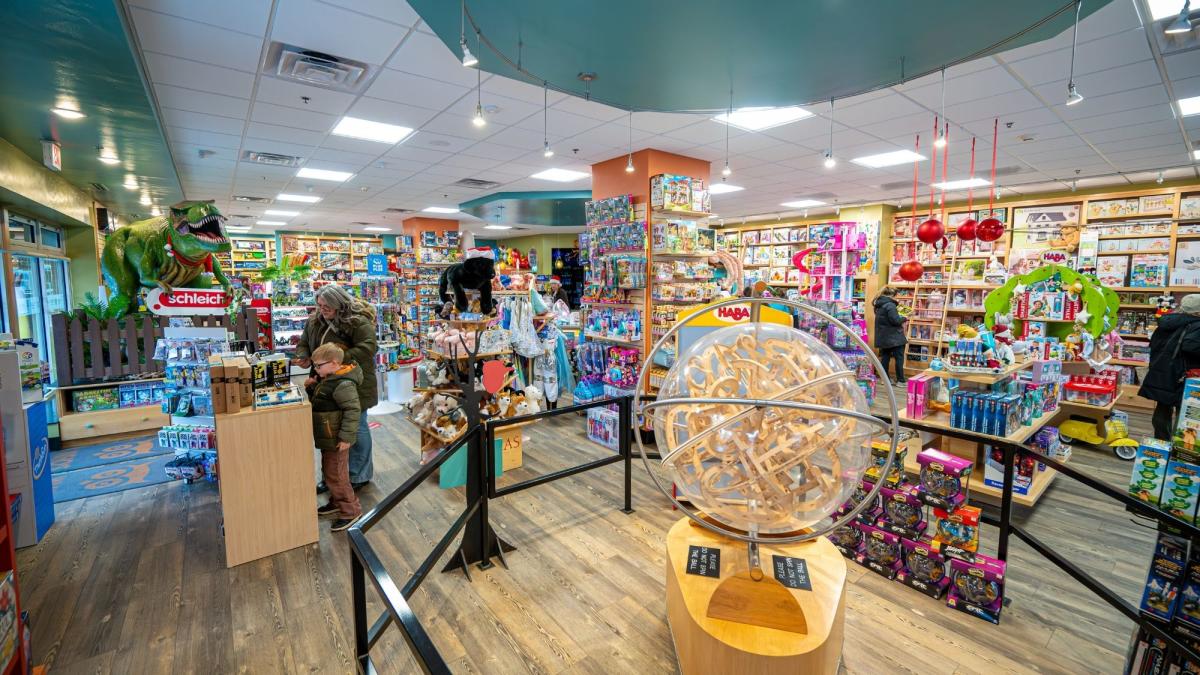A bright, colorful toy store interior filled with shelves of games, puzzles, and toys, with families browsing. A large wooden marble maze sphere sits in the foreground, while a green dinosaur display and stuffed animals add playful accents throughout the space.