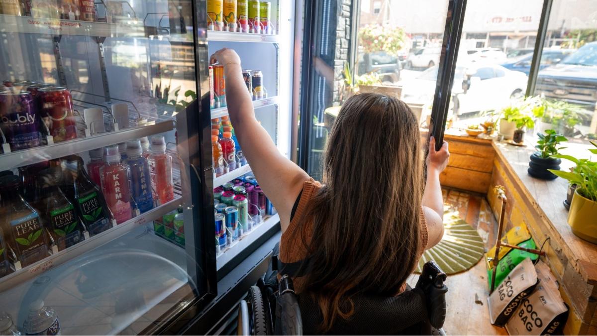 A person in a wheelchair reaches into a refrigerated display of colorful drinks, surrounded by bright sunlight and indoor plants. The scene is casual and inviting.
