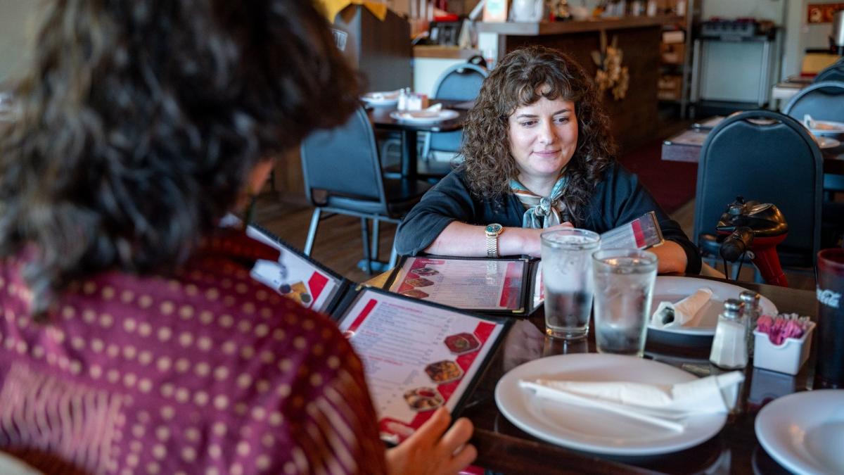 Two people sit in a cozy restaurant, looking at menus. The woman facing the camera smiles softly. The table is set with water glasses and cutlery.