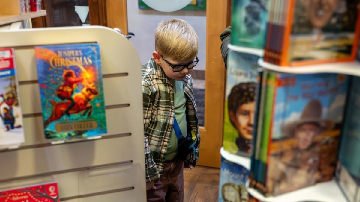 A young child wearing glasses browses between book displays in a cozy bookstore, looking down thoughtfully as colorful book covers frame the scene on both sides.