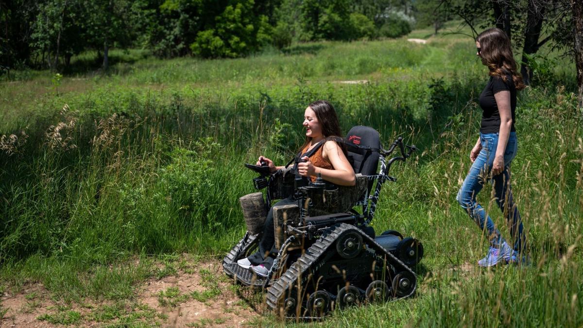 A woman in an all-terrain wheelchair, smiling, navigates a grassy path alongside another woman walking. The scene is set in a lush, green park.