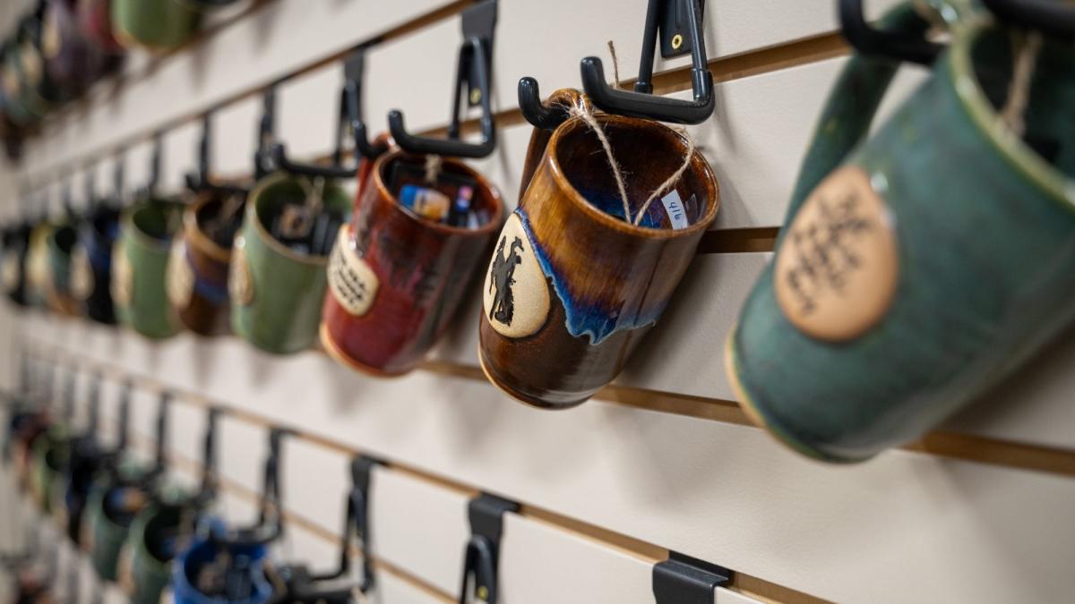 A row of colorful ceramic mugs hangs on a wall display with black hooks. The mugs feature various designs, creating a warm, artisanal vibe.