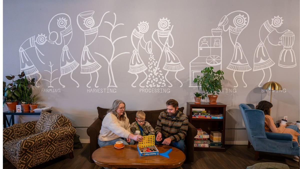 A family sits on a couch in a cozy café lounge, playing a tabletop game together at a wooden coffee table. Behind them, a large mural illustrates the coffee journey from farming and harvesting to processing and brewing, while other patrons relax in armchairs nearby.