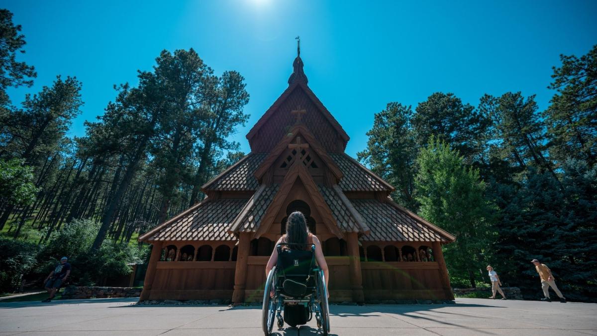 A person in a wheelchair faces a traditional wooden church amidst tall pine trees under a clear blue sky, conveying serenity and inclusion.