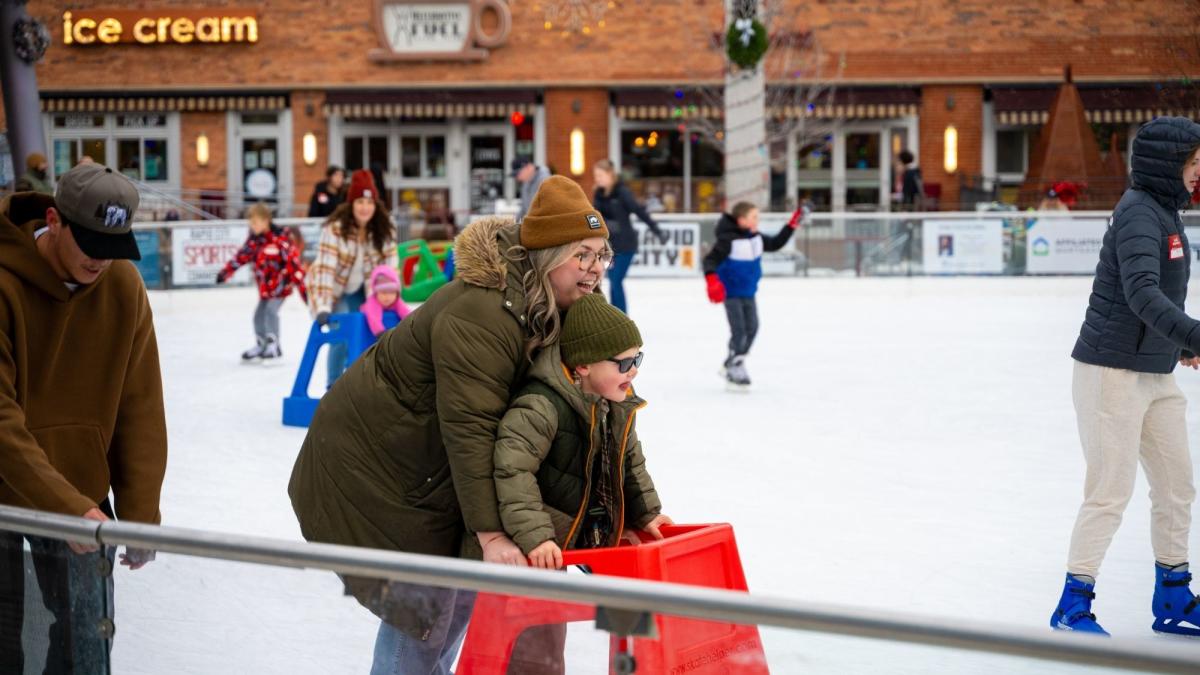 A woman and child in matching green jackets and beanies laugh while using a red skating aid on an ice rink. People and shops are in the background.