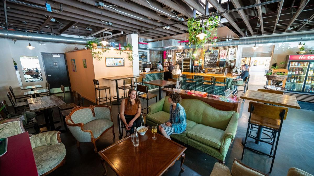 Two women sit in a cozy café on vintage furniture, engaged in conversation. The space features wooden tables, hanging plants, and a warm, inviting atmosphere.