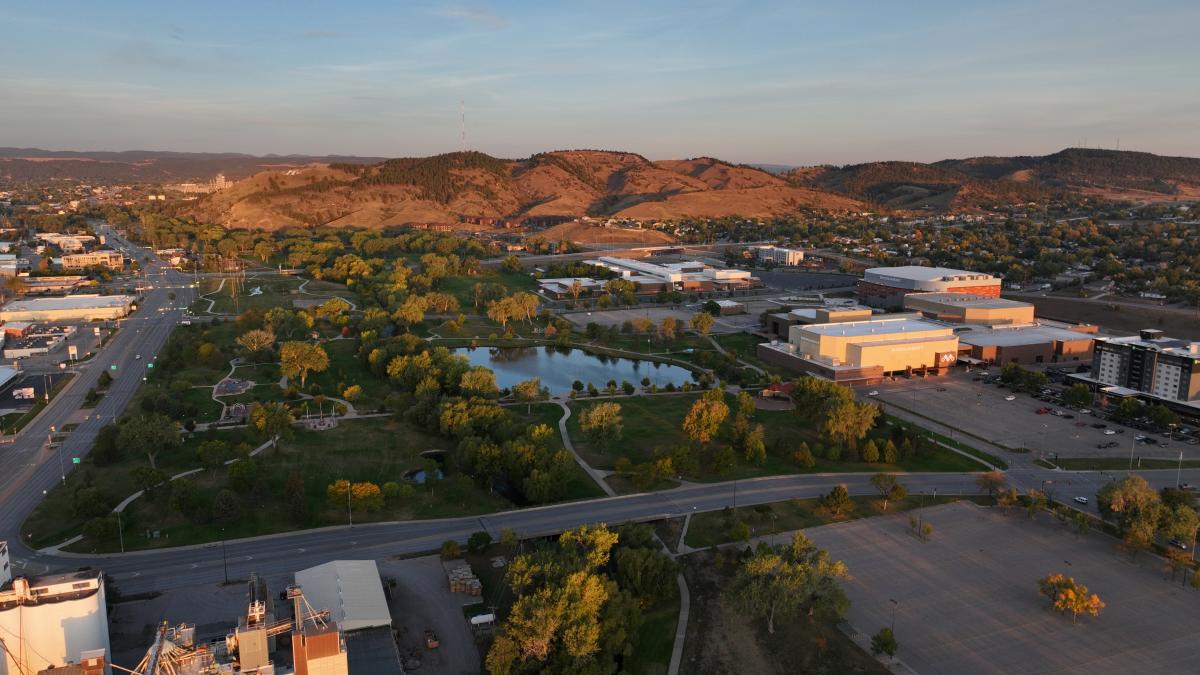 Aerial view of Memorial Park in Rapid City during golden hour, featuring winding walking trails, open green space, a small lake, and tree-lined landscapes. The Monument campus sits beside the park, while the rolling foothills of the Black Hills rise in the background.