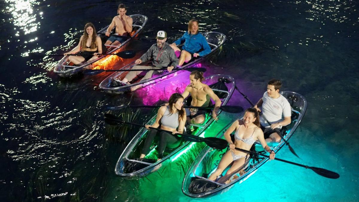 People paddling in clear kayaks at night with colored lights