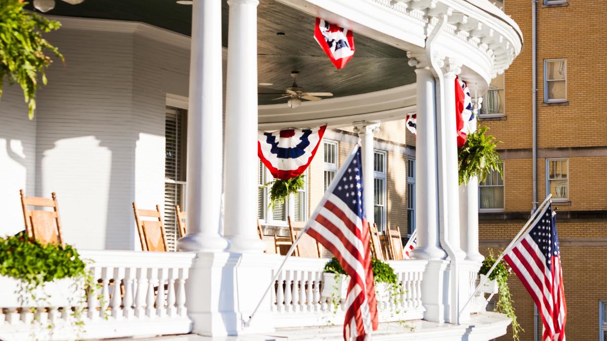 Sunny hotel veranda with white columns, rocking chairs, American flags, and patriotic bunting.